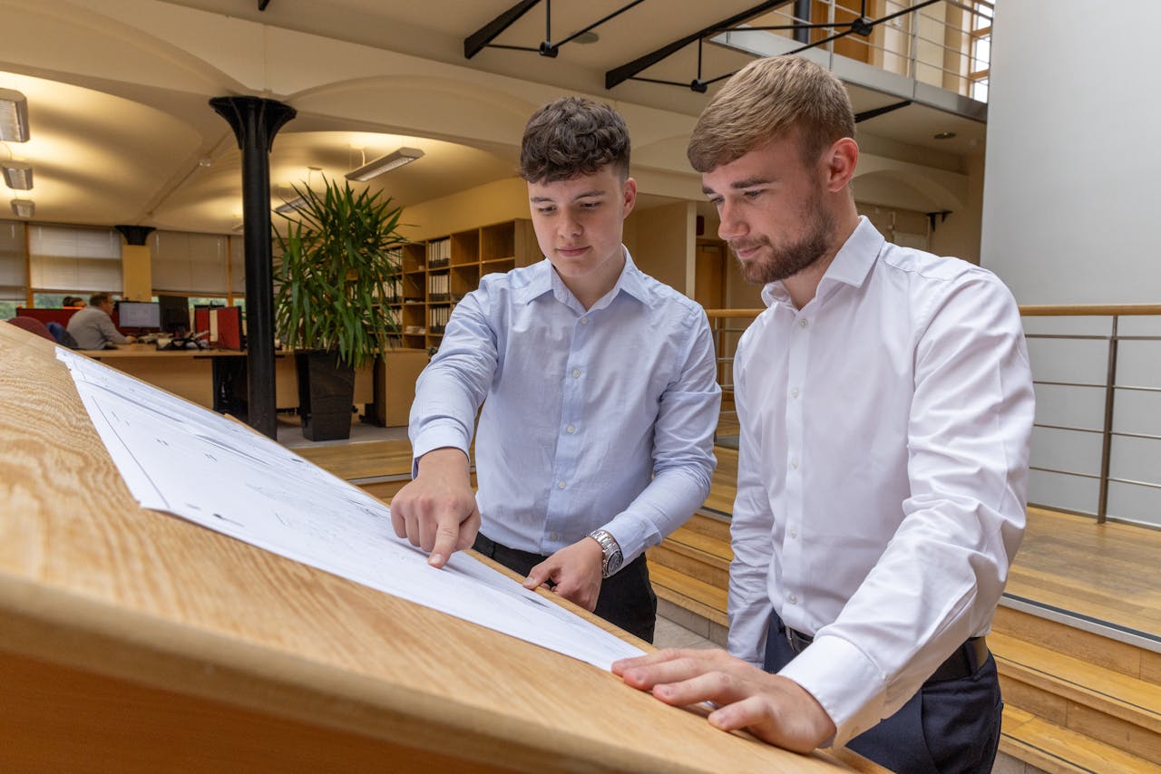 Two engineers in white shirts discuss blueprints at an office desk.